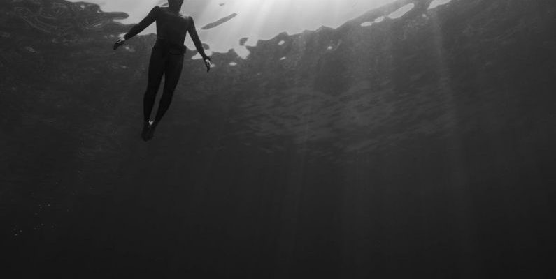 Dive Travel - Black and white of from below of scuba diver swimming in clear water in swimsuit under rays of light