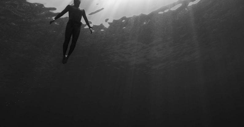 Dive Travel - Black and white of from below of scuba diver swimming in clear water in swimsuit under rays of light