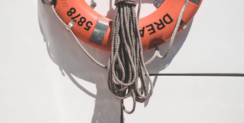 Marine Protected - Orange lifebuoy with tied mooring rope hanging on white wall of modern yacht on sunny day